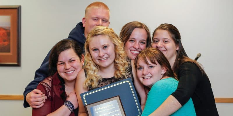 A group of students hug each other around a plaque for the A group of students hug each Lumberjack Leaders Institute.