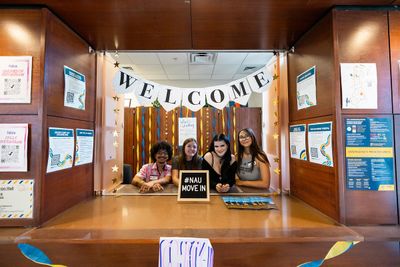 Students stand at a desk behind a sign saying #N A U Move in welcoming new students during move-in week.