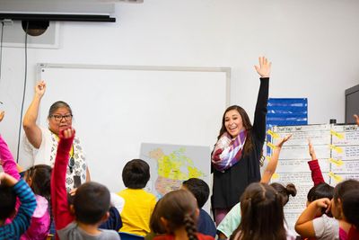 A teacher and student instructor encourages young students to raise their hands in the classroom while standing in front of a whiteboard and map of North America.