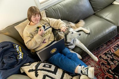 A woman does schoolwork from a laptop on her couch drinking coffee and petting a husky.