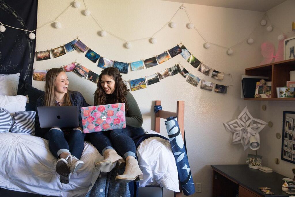 Two N A U roommates sit on a bed in their dorm room with laptops, laughing.