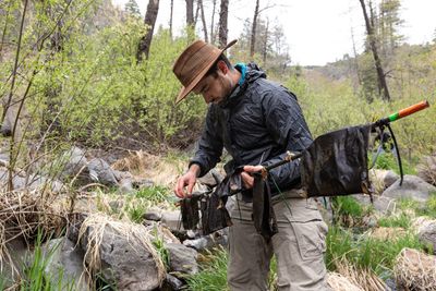 Person wearing dark jacket, hat, and pants, carrying a backpack and standing near a rocky stream in a forested area.