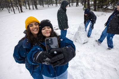 Students bundled up in jackets, beanies, and gloves, take a selfie in the snow, while others make a snowman in the background.