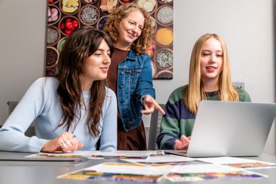 Three individuals sitting at a table, interacting with a laptop computer and some printed materials. The individuals appear to be engaged in discussion or collaborative work.