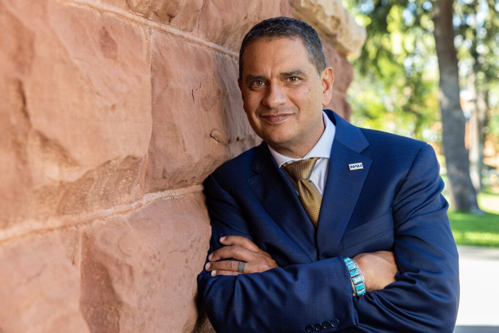 President Jose Luis Cruz Rivera in a dark blue suit leaning against a red rock wall on NAU Flagstaff campus with his arms crossed.
