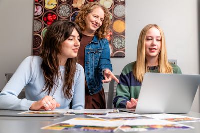 Three individuals sitting at a table, interacting with a laptop computer and some printed materials. The individuals appear to be engaged in discussion or collaborative work.