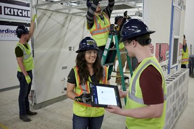 Students wearing high-vis vests, hardhats, and safety gloves work in a construction site.
