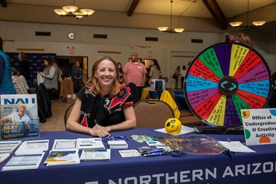 A staff member sits at a desk at a college fair next to a spinning wheel with fliers organized all over the desk.