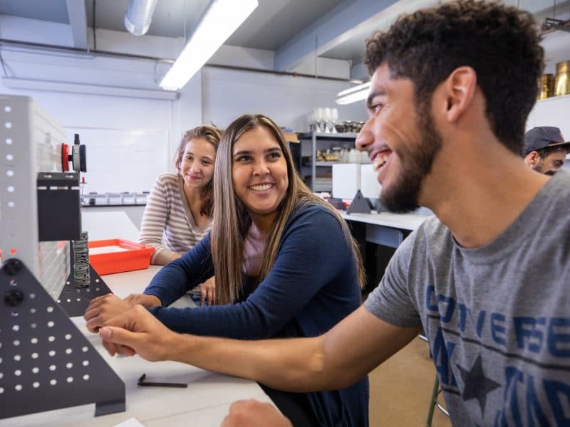 Three students sit at a bench in an engineering lab working together on an assignment