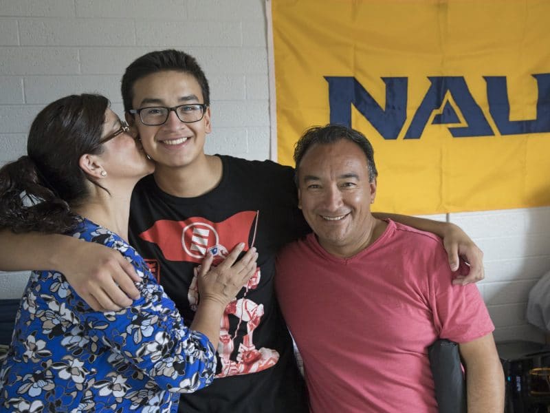 Student stands with their parents in front of NAU flag in dorm room.