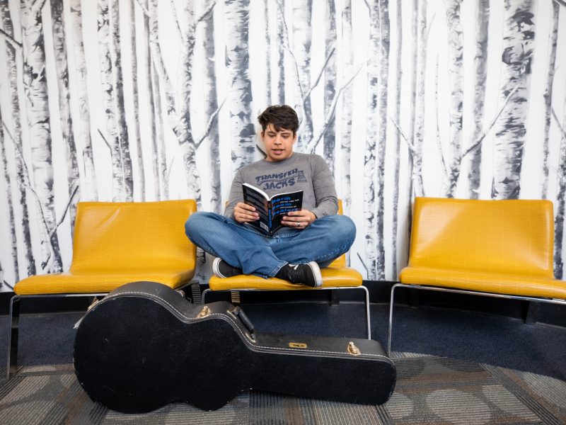 Student reads a book with guitar case at his feet.