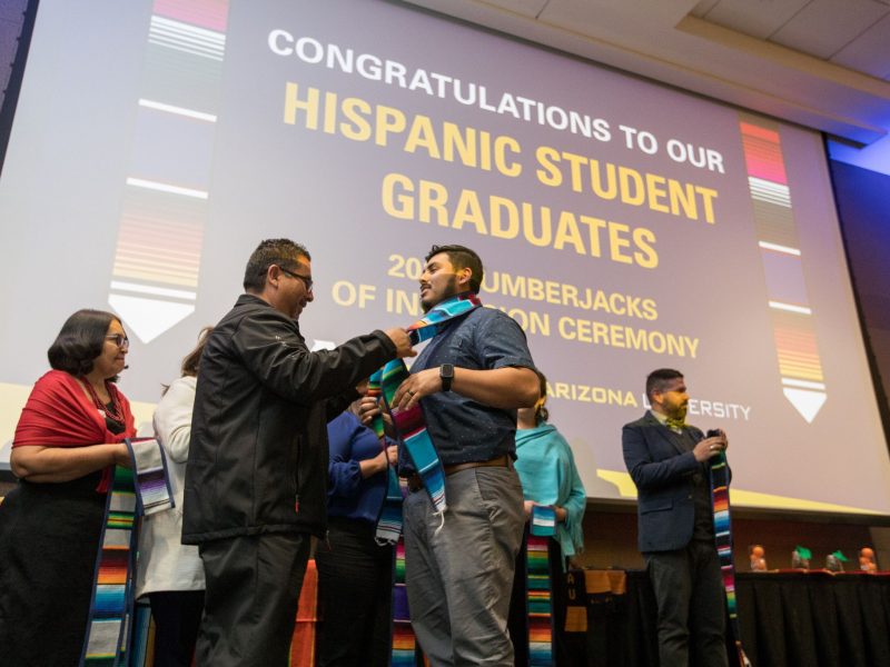 Upcoming graduate presented with a stole in front of a slide reading, 'Congratulations to our Hispanic student graduates.'