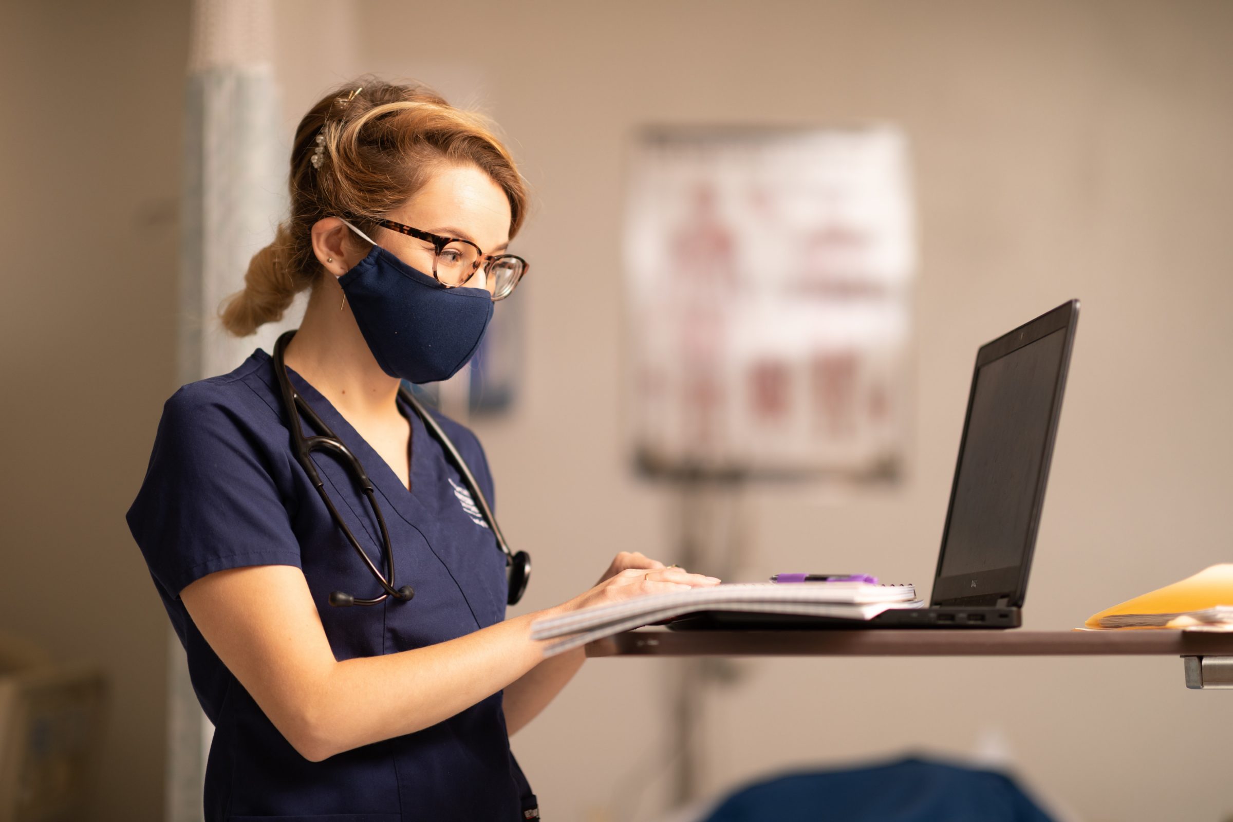 Nursing student wearing scrubs and a stethoscope takes notes on a laptop.