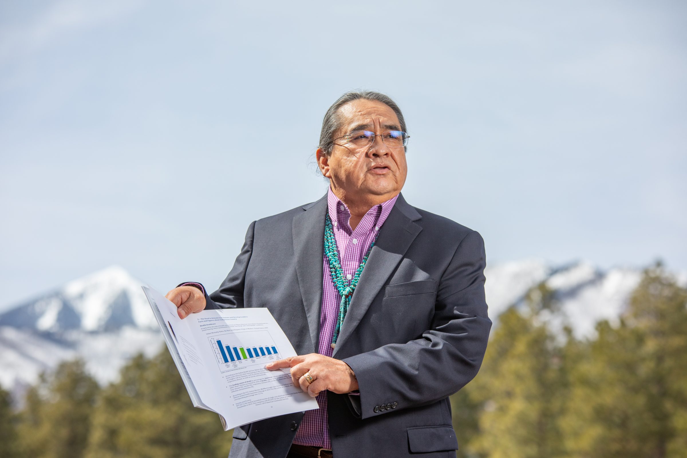 Manley Begay holds a chart and stands with mountain peaks behind him.