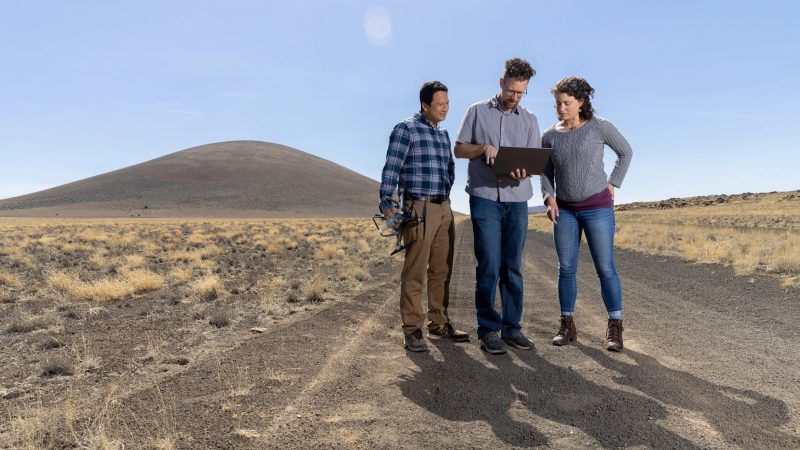 Three researchers stand outside on a dirt road looking at a laptop.