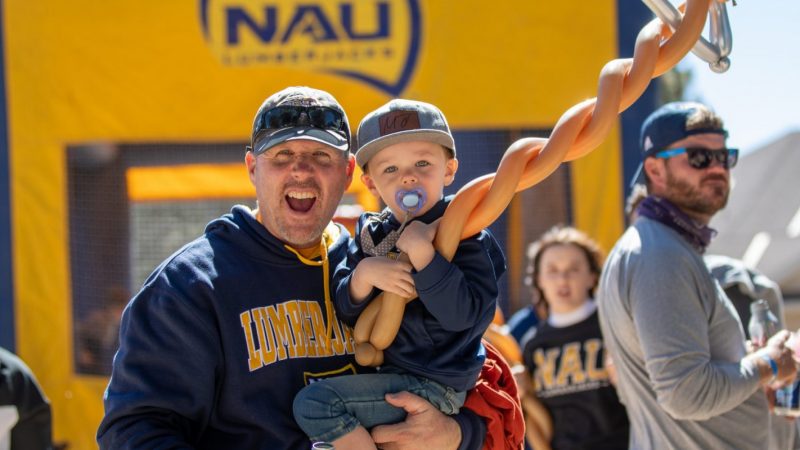 Alumni carrying his toddler son while cheering at the camera. The toddler is holding a big axe balloon.
