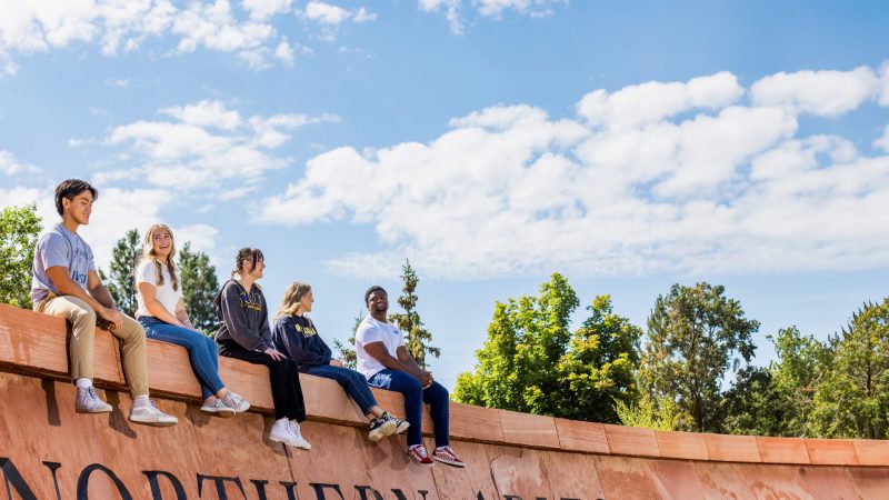 Five students sit on top of the red brick sign of N A U at Flagstaff mountain campus.