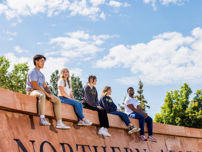 Five students sit on top of the red brick sign of N A U at Flagstaff mountain campus.