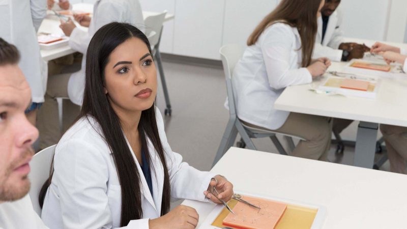 A student in a lab coat practices suturing.