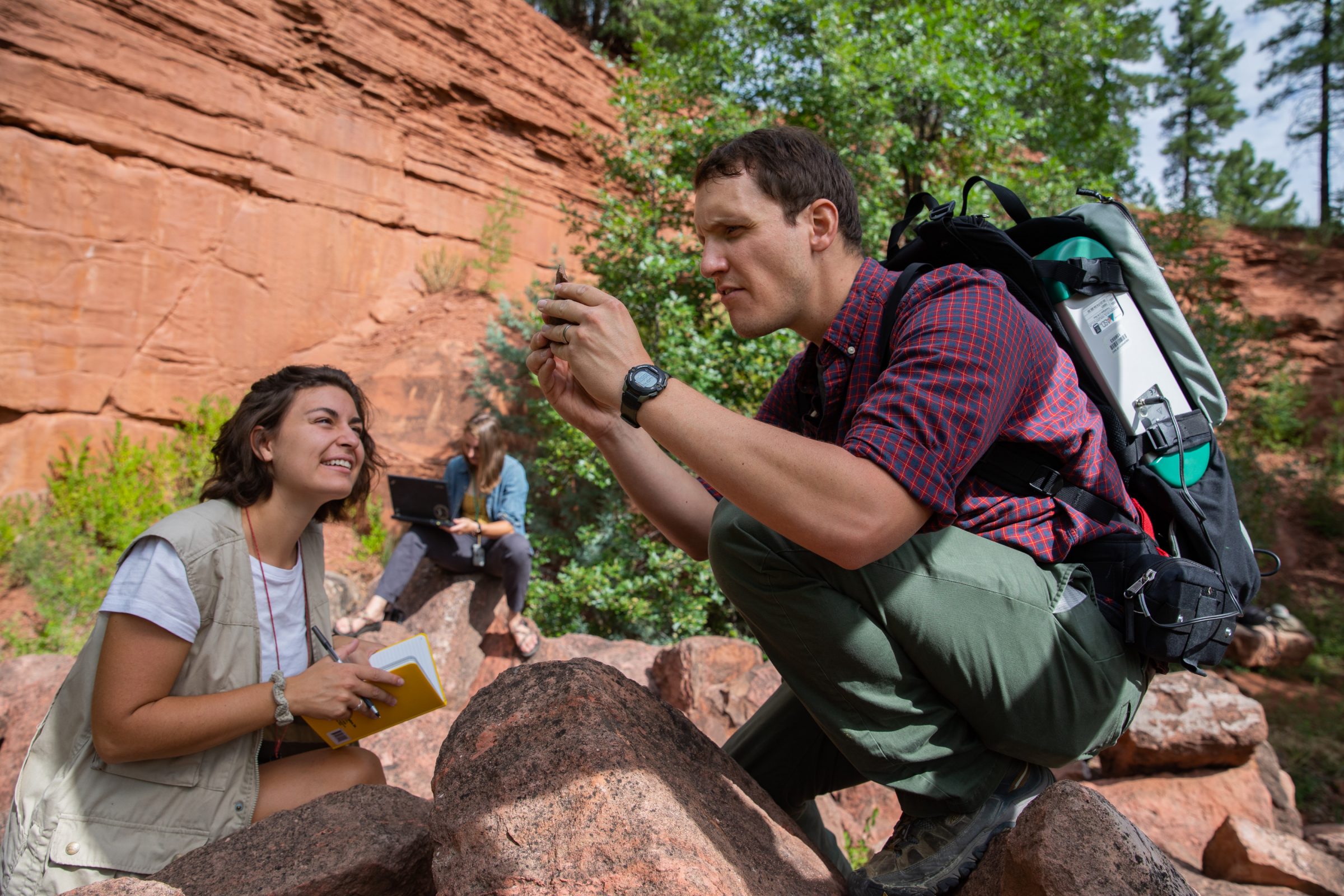 Two students outside while one is perched on a rock looking at a phone.