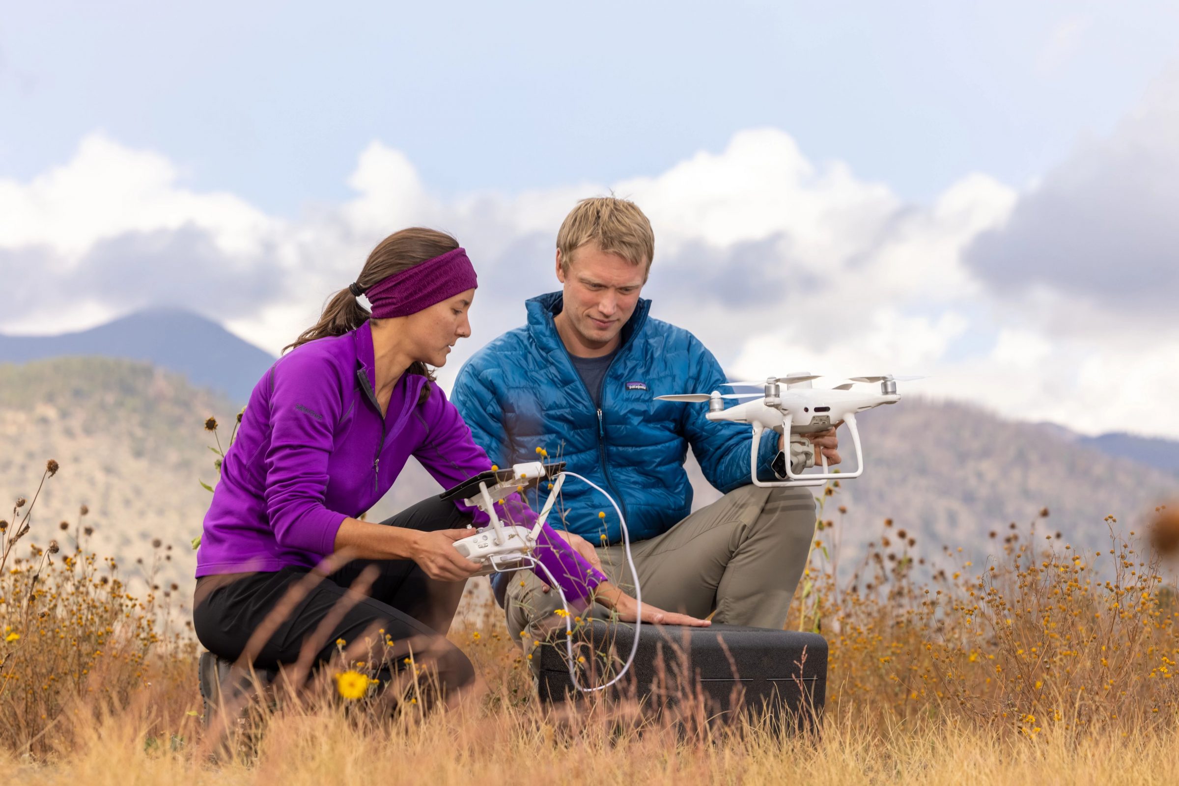 Two people are crouching down in a field, one is holding a drone and the other is holding the remote.