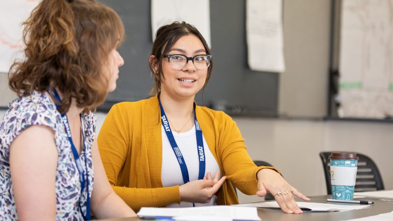 Two students are sitting in a classroom while discussing a project.