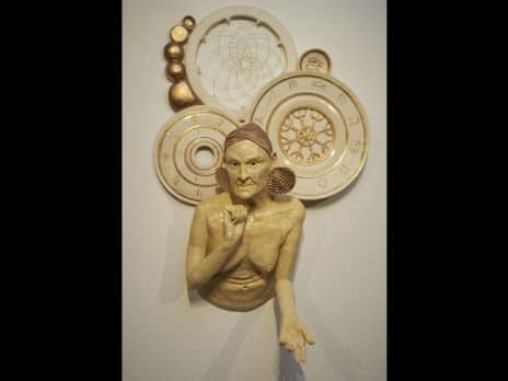 Wall-mounted ceramic sculpture of an older woman's upper body. She's wearing large earrings and there are large discs with zodiac symbols and other ancient symbols behind her. Sculpture by student Hailey Campanella