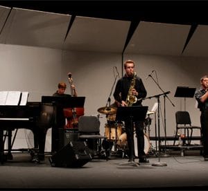 student plays saxophone on stage while student plays the cello behind him