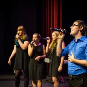three female students and one male student sing into microphones on stage