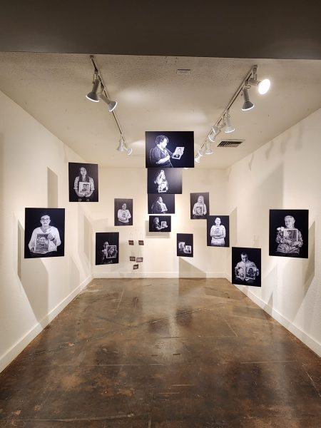 The image depicts a gallery with white walls with black and white photographs hanging from the ceiling depicting family members of people murdered in Mexico holding photos of the victims.