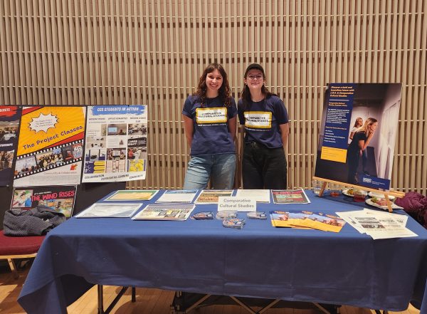 The image shows CCS majors Joce Dolezal and Ella Conner wearing blue and gold Comparative Cultural Studies t-shirts standing behind a table with flyers and various posters behind them.