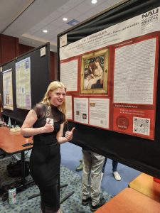 A woman with long blond hair in a black outfit stands in front of a red research poster.