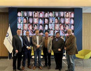 A group of men in suits post before a projector screen after a panel.