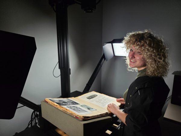 Alex Williams looks over her shoulder at camera from an open book of historical photos she has in front of her. A bright light is directed in front of her at the book.