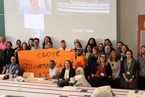 A group of people pose with a large orange sign stating "Close Guantanamo" in front of a projector screen.