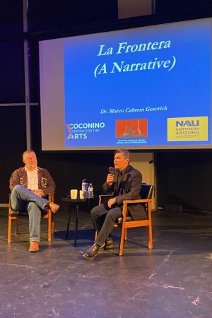 The image shows two men seated on a stage in front of a screen which reads La Frontera (A Narrative), the title of the presentation by Dr. Marco Cabrera Geserick. 