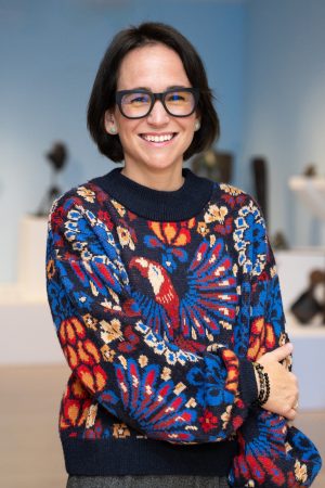 This image shows a woman with dark short hair and glasses wearing a brightly patterned sweater in an art gallery.