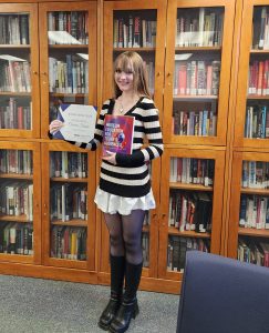 A student in a black and white sweater and long hair holds a certificate and a book.