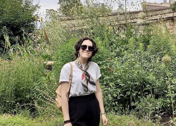 This image is a photograph of CCS alum Emily Morgan. A woman stands in a green garden outdoors with short curly hair and sunglasses.