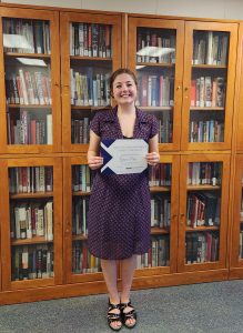 A woman in a purple dress stands in front of a bookcase holding a certificate.
