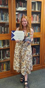 A student in a flowered dress holds a book and a certificate. 