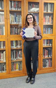 A student wears glasses and a purple shirt in front of a bookcase holding a certificate.