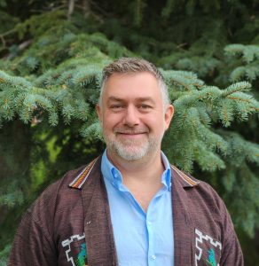 The image shows a man with light hair and beard with a blue and brown shirt in front of a pine tree.