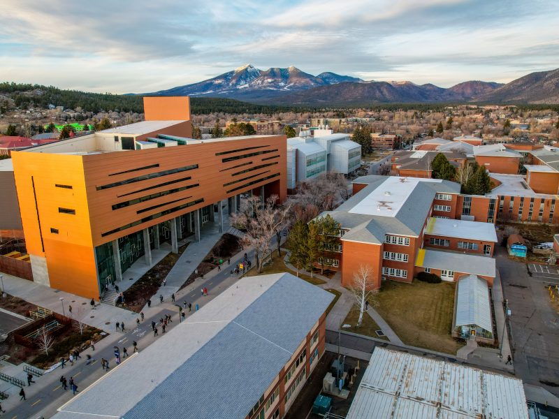 Photo taken from high in the sky, NAU's science building is captured from above, with the city of Flagstaff and San Fransisco Peaks in the background.