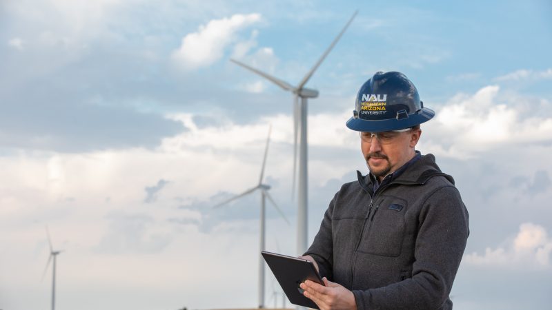 A man wearing a NAU hardhat reads a tablet with large wind turbines in the background.
