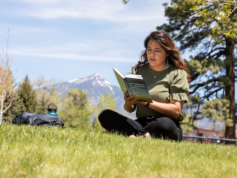A student reads a book in the grass with the San Fransisco Peaks in the background.