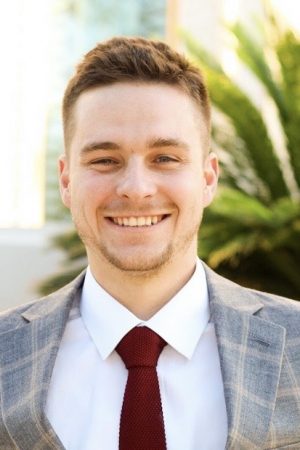 A photo of Tanner Nelson who is wearing a sportcoat, red tie, and white shirt, has short brown hair, and is against a natural background.