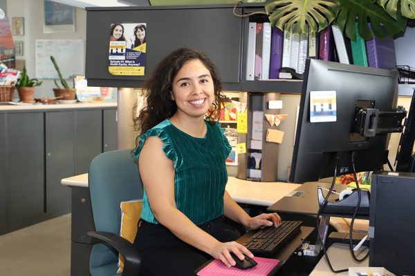 Alexandra Samarron Longorio sits at her desk in the ARD building of Northern Arizona University. She has long, dark, curly hair, is smiling, and is wearing a green velvet short-sleeved shirt.