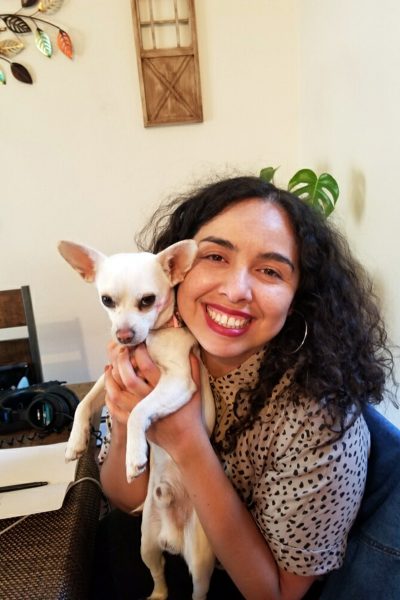 A portrait of Alexandra Samarron Longorio sitting a dining table. She is smiling and holding her white chihuahua, Randall. She is has dark, curly hair parted in the middle and is wearing silver, hoop earrings.