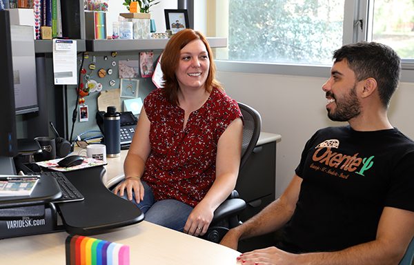 Amanda Pollitt, sits in a black office chair behind her desk next to one of her students. She is smiling and has red, shoulder-length hair and is wearing a sleeveless blouse with small flowers and is wearing jeans. The student has short dark hair and a beard and mustache. They are wearing a black T-shirt with the word Oxente on the chest with a green cactus.